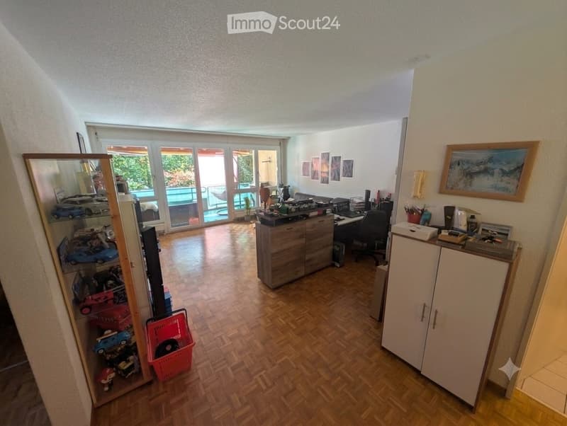 Spacious room with wooden flooring, large sliding glass doors leading to a balcony, a cabinet, desk, chair, mirror, framed picture, and a basket with toys.