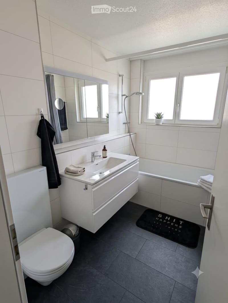 Modern bathroom with white tiles, a large mirror, a sink with a modern faucet, a toilet, a bathtub, and a small window with a plant on the sill. A black towel hangs on the wall, and a bath mat is placed in front of the bathtub.