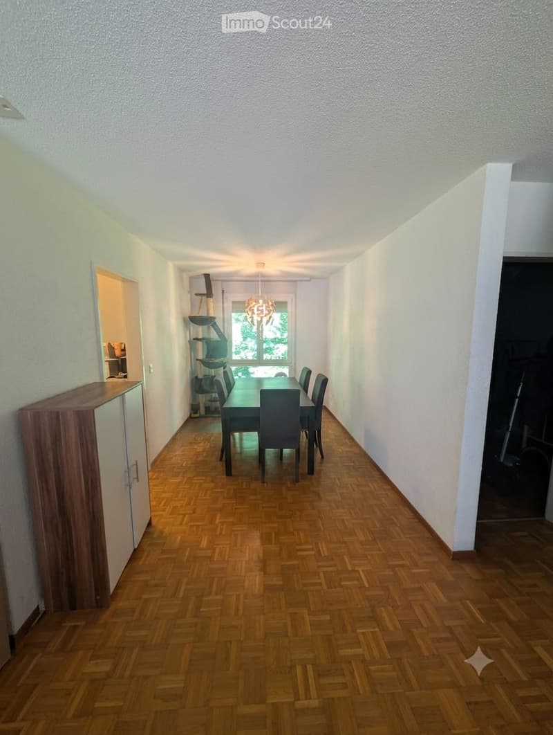 Dining room with wooden herringbone flooring, white walls, and a dining table with six chairs. A cabinet is visible on the left side, and a window with a view of greenery is at the far end.