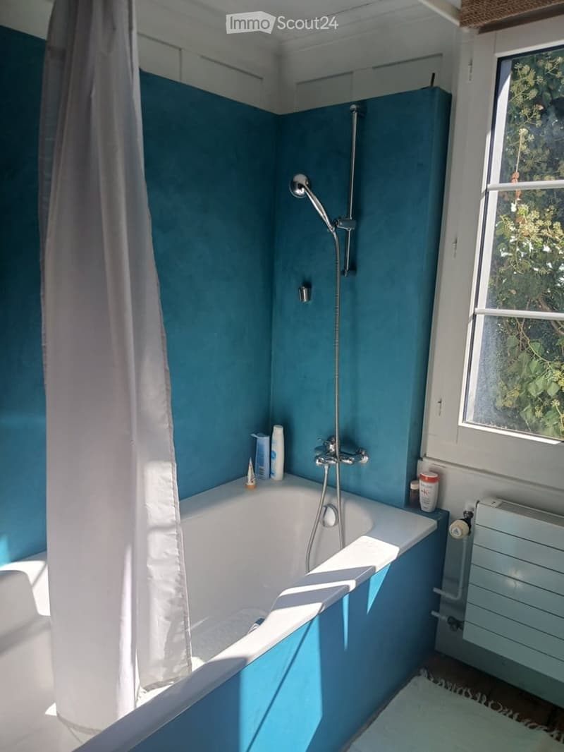 Bathroom with blue walls, white bathtub, showerhead, radiator, and various toiletries on the edge of the tub.