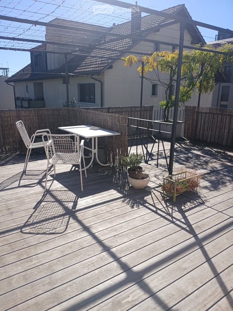 Wooden deck terrace with white metal table and chairs, potted plants, and a wooden fence, with a house visible in the background.