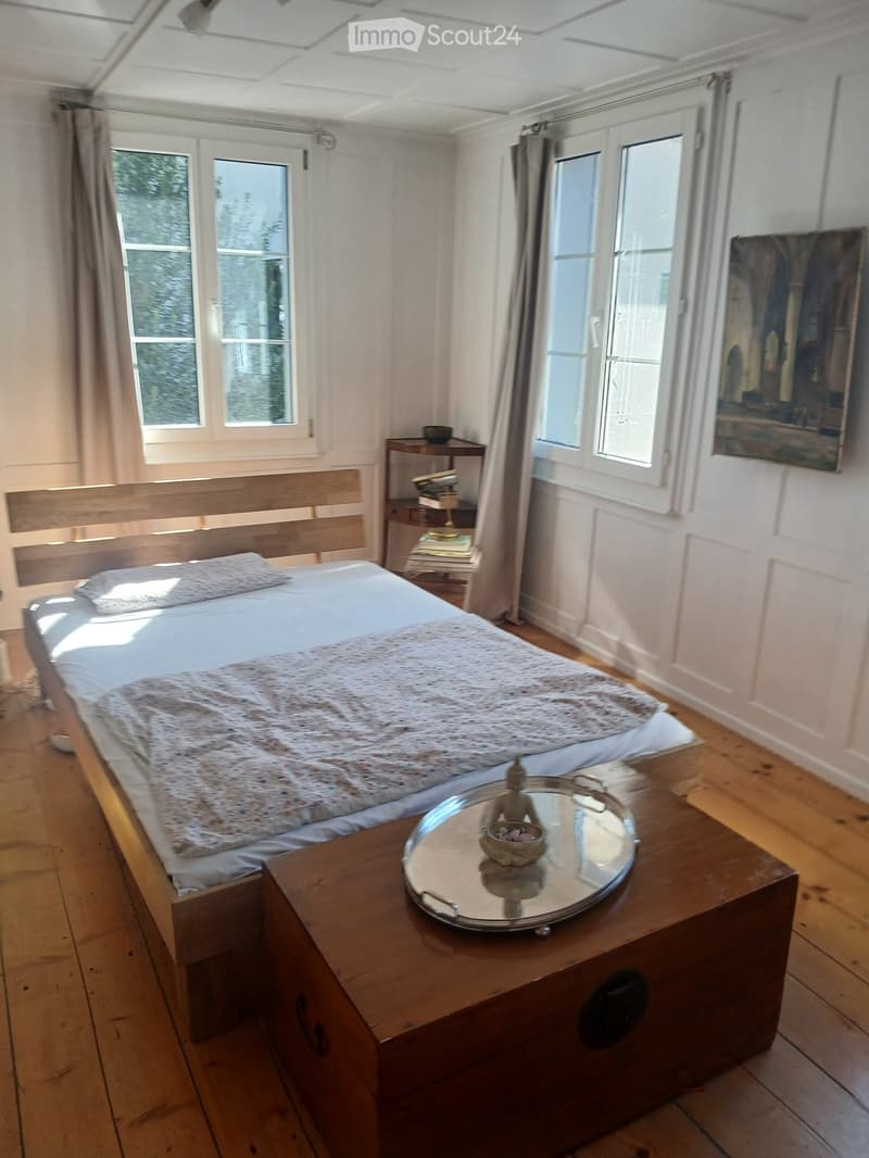 Cozy bedroom with wooden floors, large windows, a neatly made bed with a fur throw, a nightstand with a lamp, a small table with a decorative tray and a figurine, and a painting on the wall.