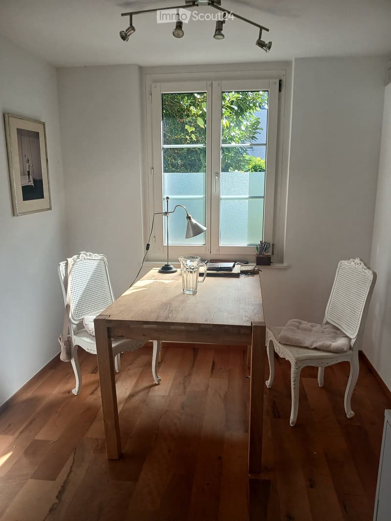 Bright room with wooden floor, large window, table with two chairs, lamp, and a cup on the table.