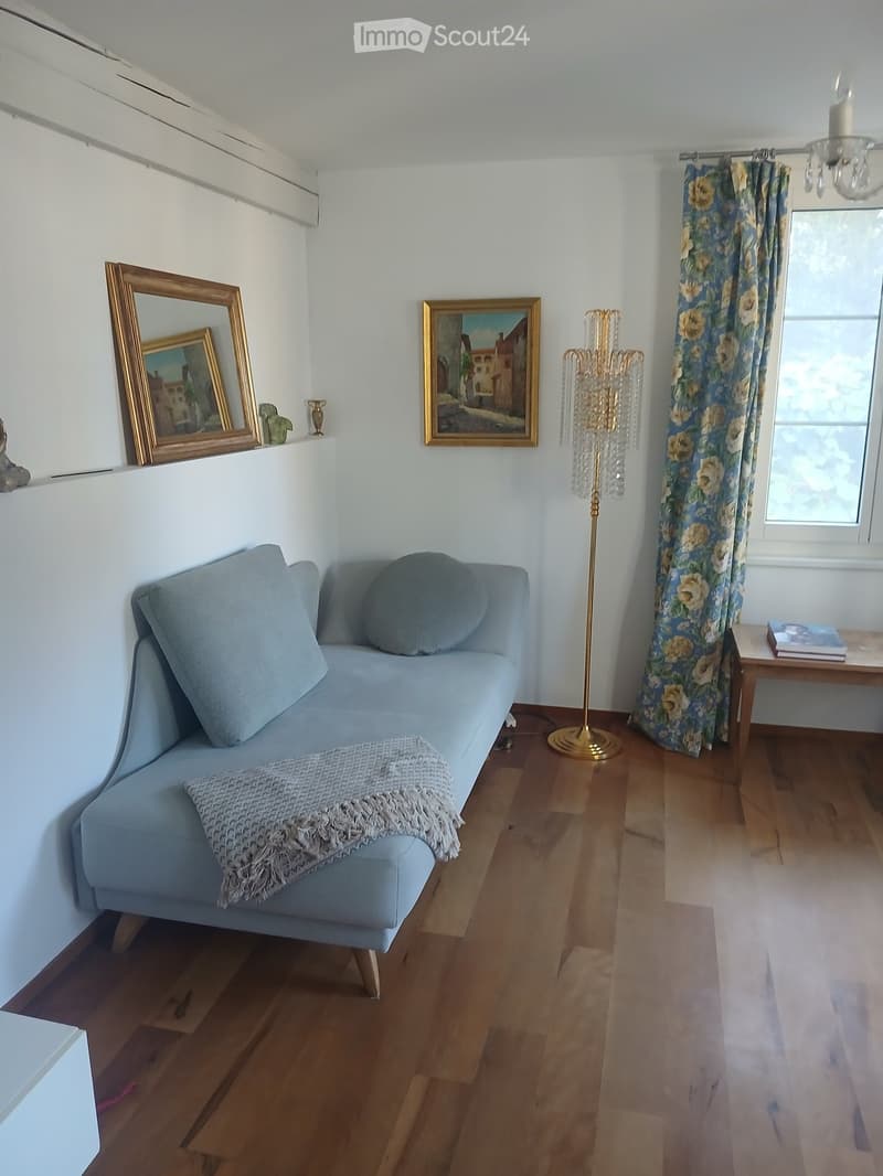 A modern living room with a light blue sofa, a wooden coffee table, a tall floor lamp, and floral curtains. The room features wooden flooring and white walls with decorative picture frames.