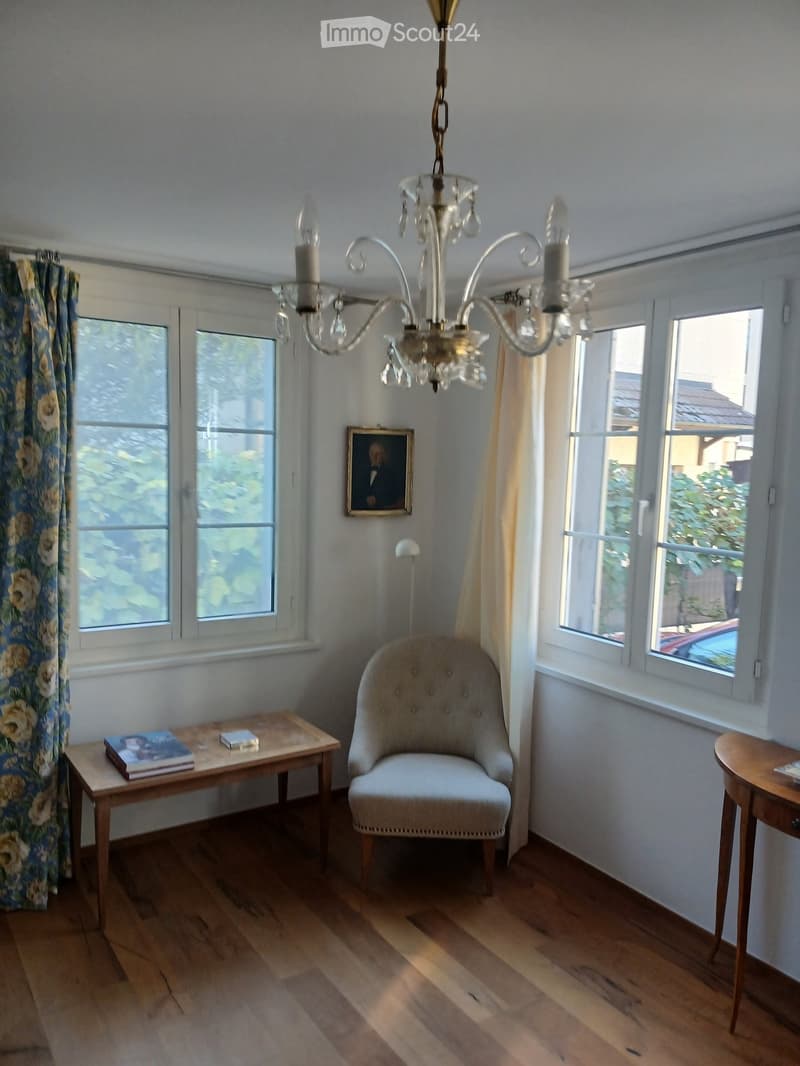 Bright room with wooden flooring, large windows, a chandelier, a single armchair, a small table with books, and a framed picture on the wall.