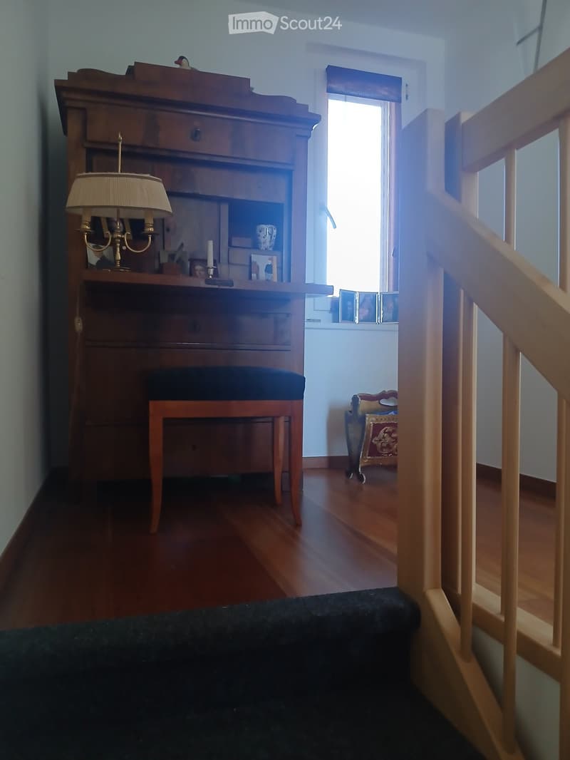 A small, well-lit room with wooden flooring, a wooden table with a lamp, a stool, and a small bench. The room has a window with a view of another room.