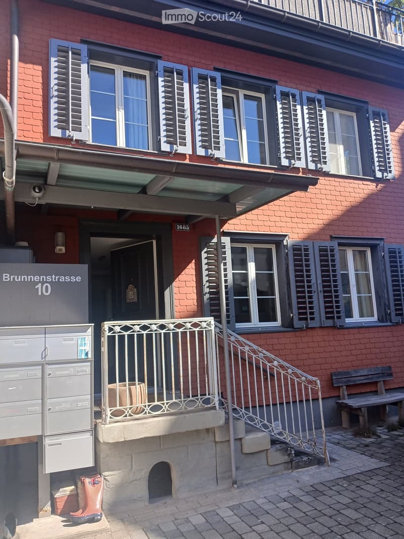 Two-story red brick building with multiple windows, shutters, and a small balcony with metal railing. The entrance has a staircase leading up to a door, and there is a mailbox and a bench nearby.