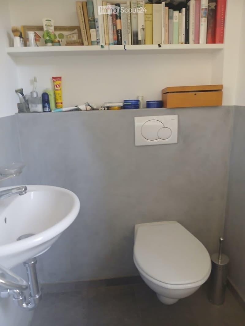 Modern bathroom with a white sink, toilet, and a shelf above holding books and toiletries. The walls are grey, and a light switch is visible.