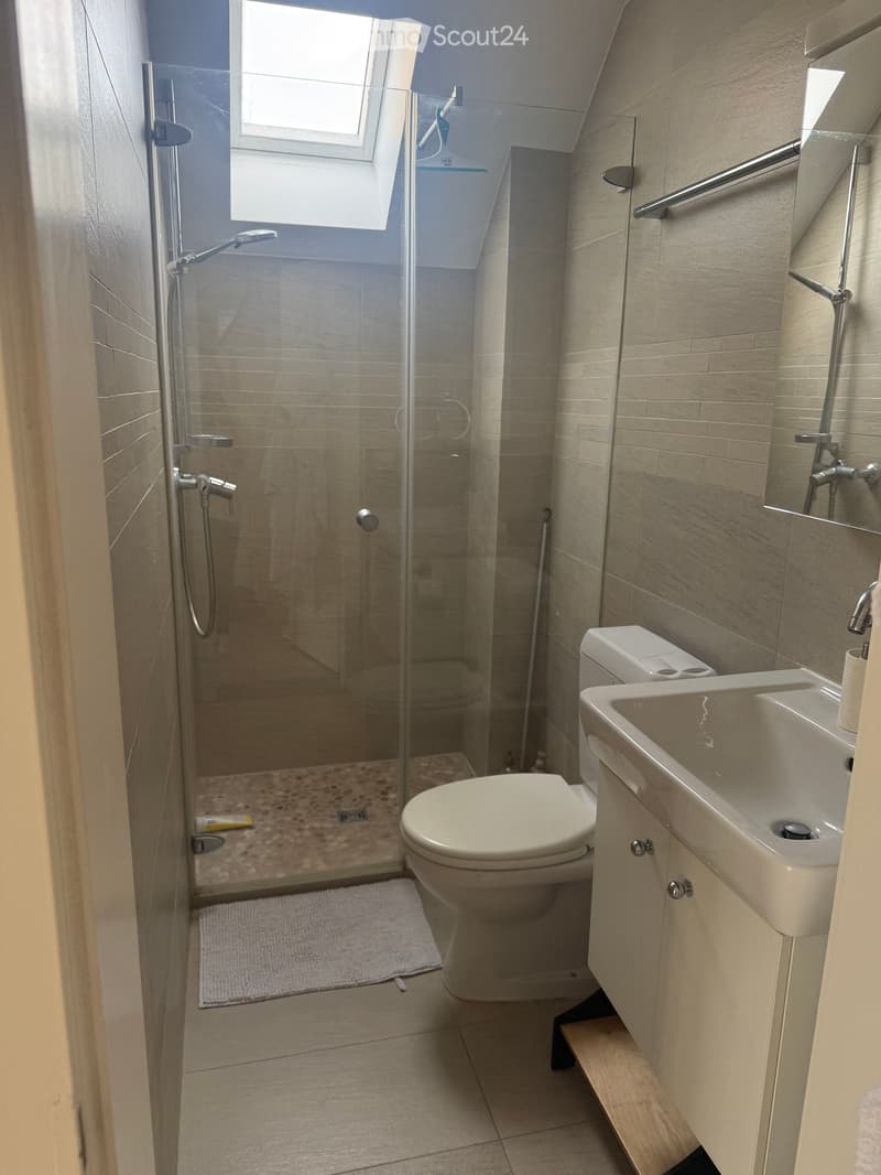 Modern bathroom with a glass-enclosed shower, toilet, sink, and a small wooden step stool. The walls are tiled in a light beige color, and the floor has a tiled pattern.