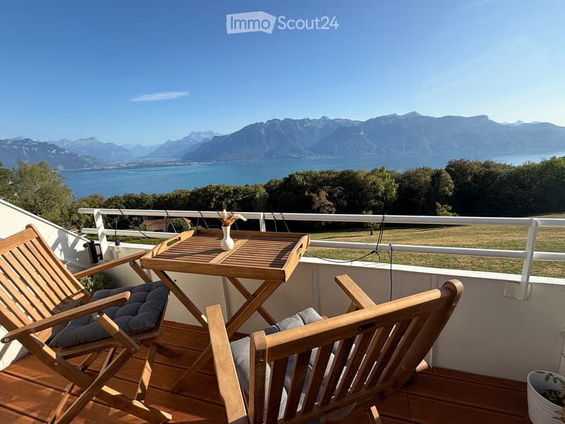 Balcony with wooden table, two wooden chairs, and a vase, overlooking a scenic lake and mountain view.
