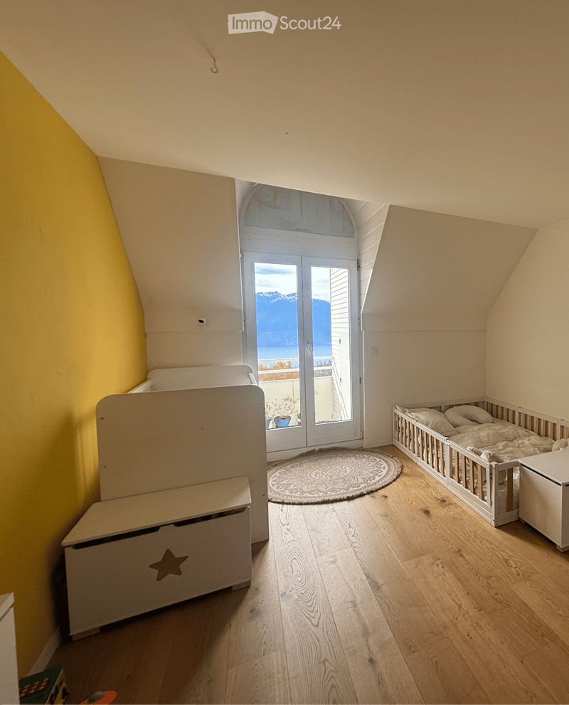 Bright, slanted-ceiling bedroom with wooden floors, a crib, a bench, and a round rug near a large window offering a scenic view.