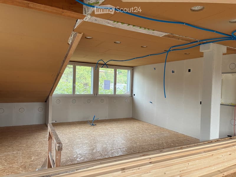 Empty attic space with exposed wooden beams, insulation, and unfinished walls. Features large windows and visible construction materials.