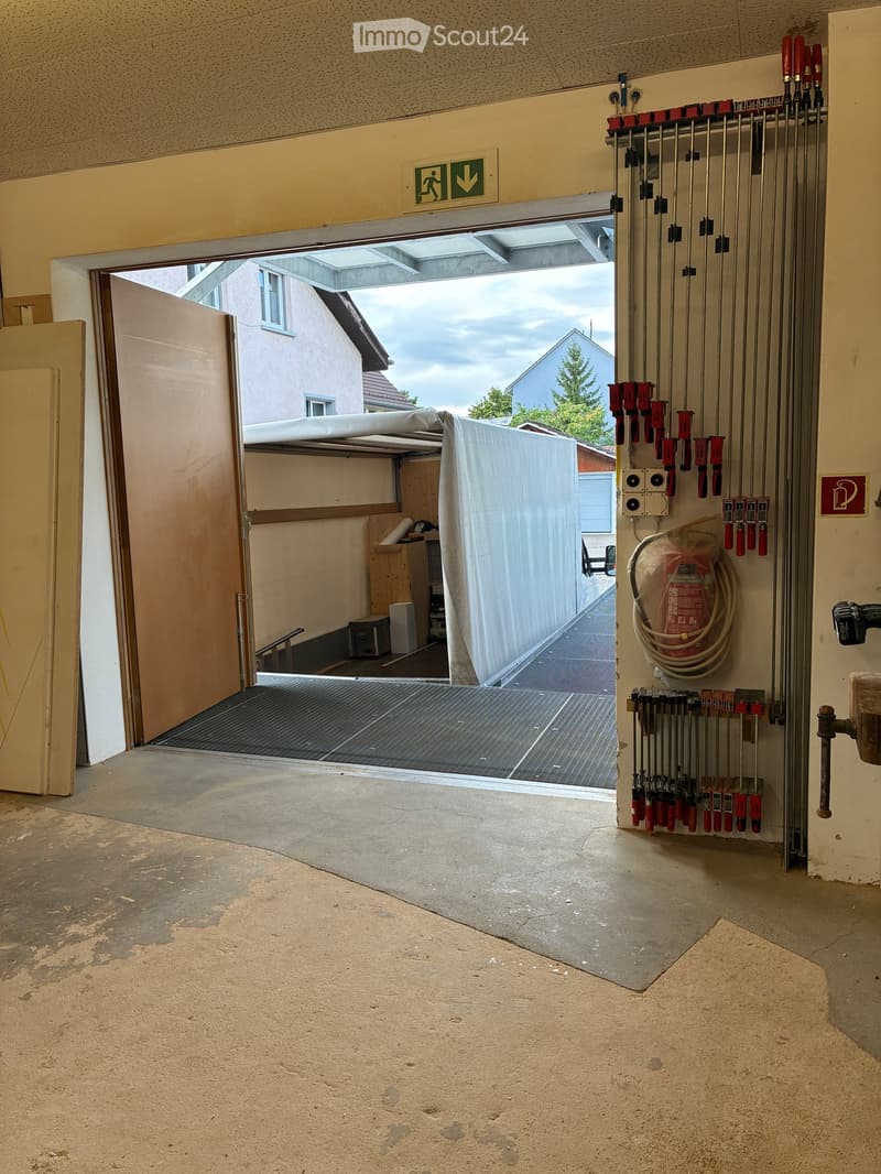 Empty garage with concrete floor, open door leading to outdoor area, tools and equipment hanging on wall, and a covered vehicle outside.