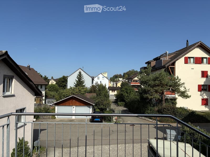 Residential neighborhood with multiple houses, trees, and a parked car visible from a balcony perspective.