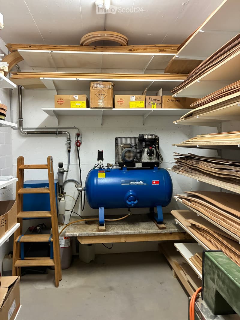 Workshop with a large air compressor on a workbench, wooden ladder, shelves filled with cardboard boxes and wooden planks.