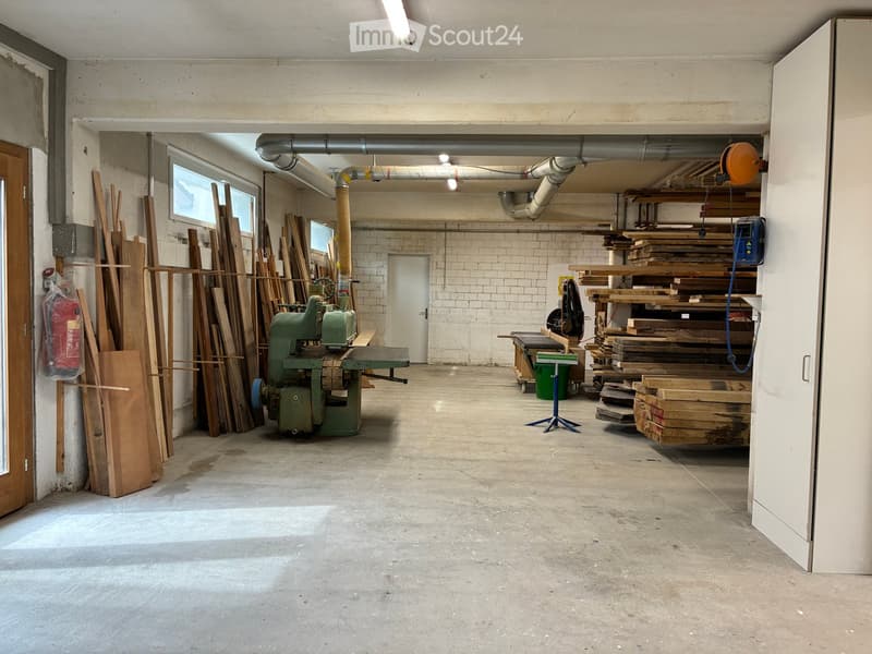 Empty workshop with concrete floor, exposed brick walls, and large windows. Contains wooden planks, a green woodworking machine, and stacked lumber.