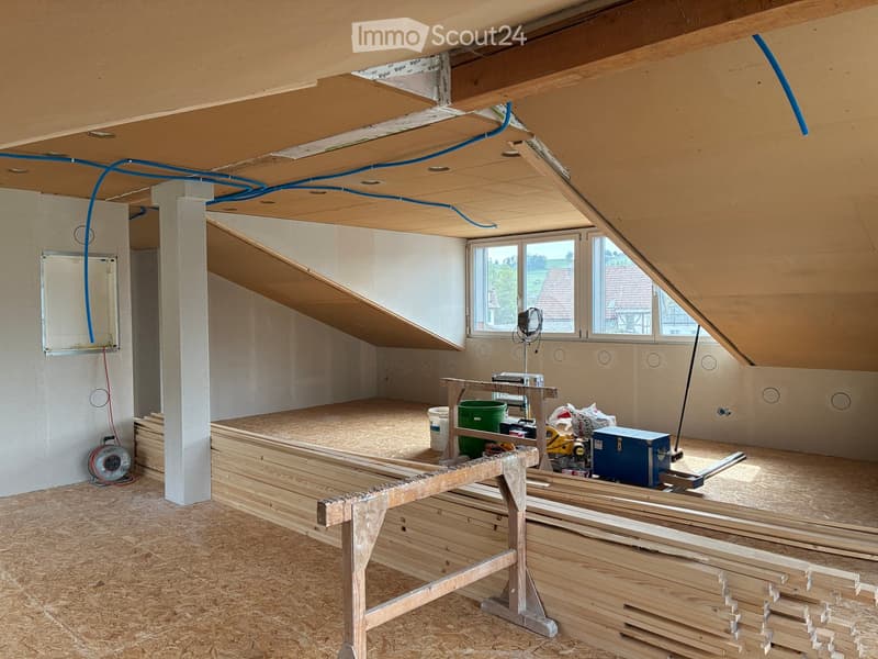 Empty attic space with exposed wooden beams, insulation, and construction materials. Windows provide natural light, and the floor is covered with plywood.