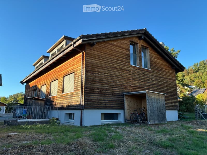 Two-story wooden house with multiple windows, a small balcony, and a bicycle parked in a wooden shed.