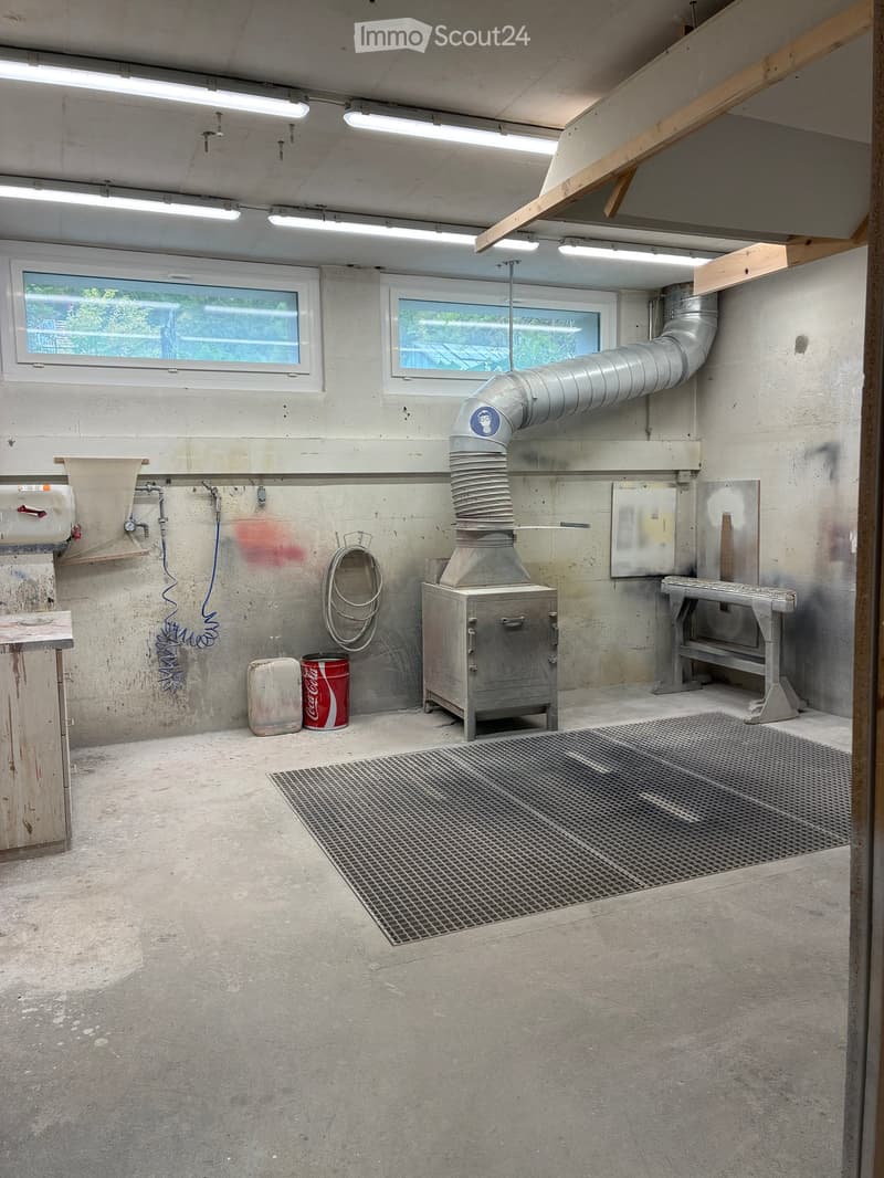 Empty room with concrete walls and floor, metal mesh floor section, ventilation duct, windows, and industrial equipment.