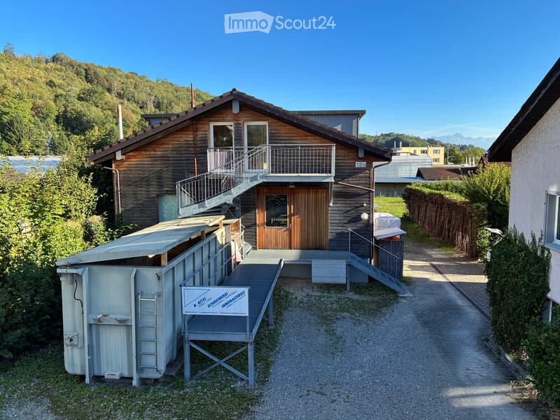 Two-story wooden house with a balcony, staircase, and a small shed with a ladder. The house is surrounded by greenery and has a clear view of mountains in the background.