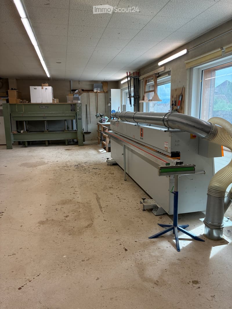 Workshop with industrial machinery, workbench, storage boxes, and tools. Natural light from windows illuminates the space.