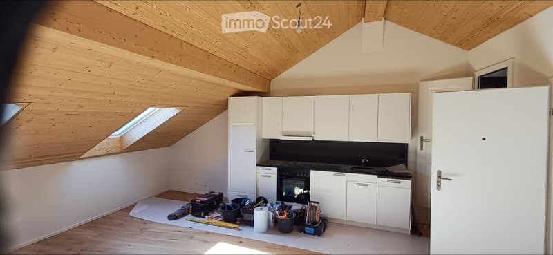 Modern kitchen with white cabinets, black countertop, and an oven. The room features wooden flooring and a skylight window, with construction materials and tools scattered on the floor.
