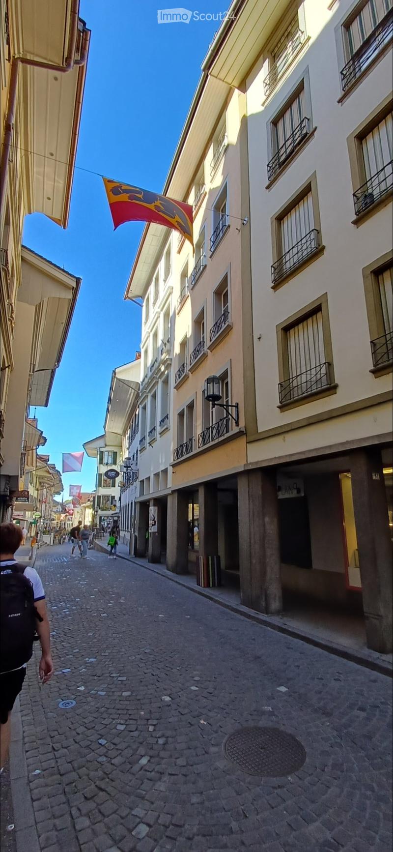 A narrow cobblestone street lined with multi-story buildings featuring balconies and awnings. A flag is visible above one of the buildings.