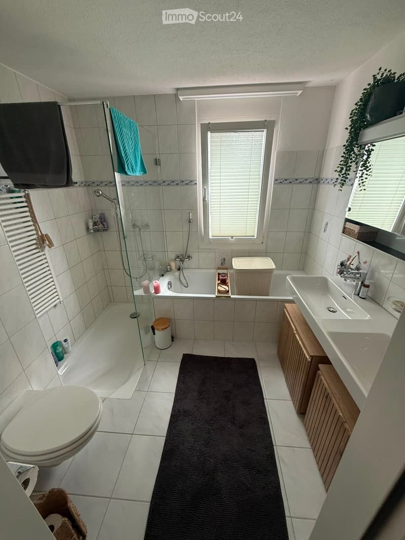 Modern bathroom with a bathtub, toilet, sink, and shower area. Features include a dark bathmat, wooden cabinets, and a window with blinds.