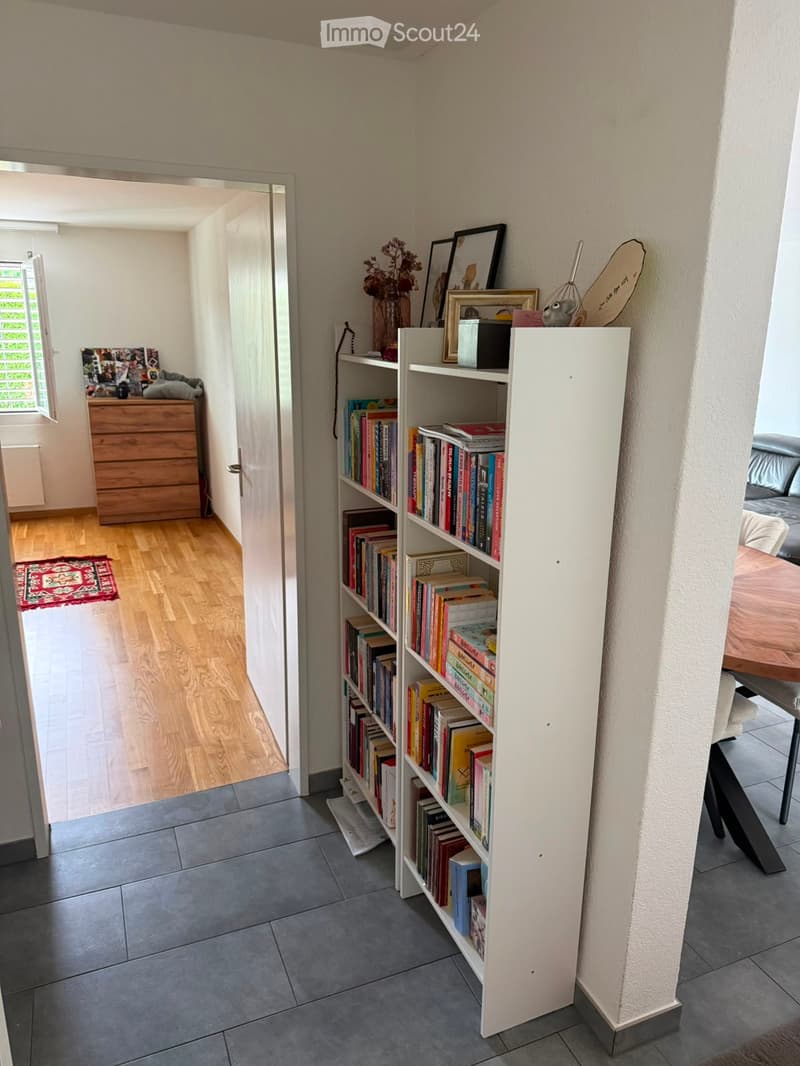 A well-lit hallway with a bookshelf filled with books, a wooden dresser, and a small rug on the floor. The hallway leads to another room with a visible bed and table.