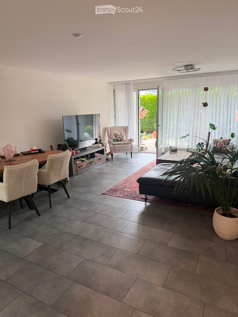 Open-plan living and dining area with grey tiled flooring, large windows, and a sliding glass door leading to a balcony. Features a black leather sofa, white armchair, wooden dining table with chairs, TV stand, and decorative plants.