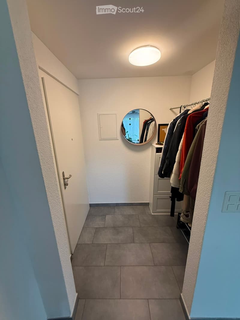 A narrow hallway with tiled flooring, white walls, a round mirror, a coat rack with several jackets, and a small shelf with a framed picture. The hallway has a closed door on the left and a ceiling light fixture.
