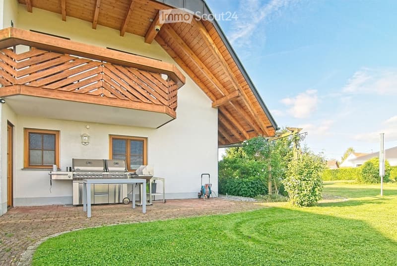 Outdoor area with a modern barbecue setup, including a grill and a table, surrounded by a well-maintained lawn and a wooden deck with a slanted roof.