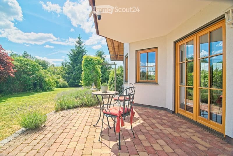 Brick-paved balcony with a small round table and two chairs, surrounded by greenery and a view of a lawn.