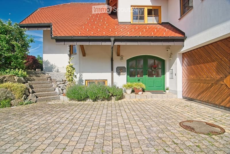 Two-story house with red-tiled roof, white walls, green front door, and wooden garage door. Paved driveway and small garden area with plants and steps leading up to the house.