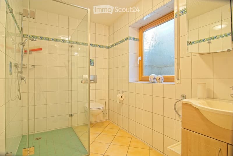 Modern bathroom with white tiled walls, a glass-enclosed shower, a toilet, a sink with a wooden cabinet, and a window with frosted glass. The floor is tiled in beige and green.