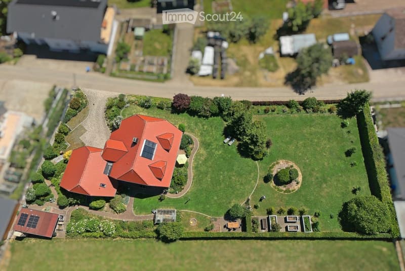 A two-story house with a red-tiled roof, surrounded by a well-maintained garden with pathways, trees, and a circular flower bed.