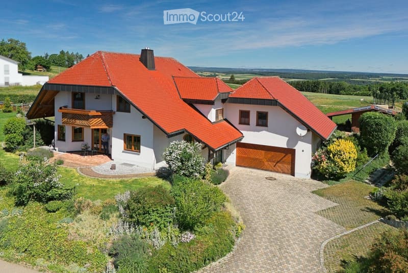 A two-story house with a red-tiled roof, white exterior walls, and a wooden garage. The house features multiple windows, a balcony, and a well-maintained garden with a paved driveway.