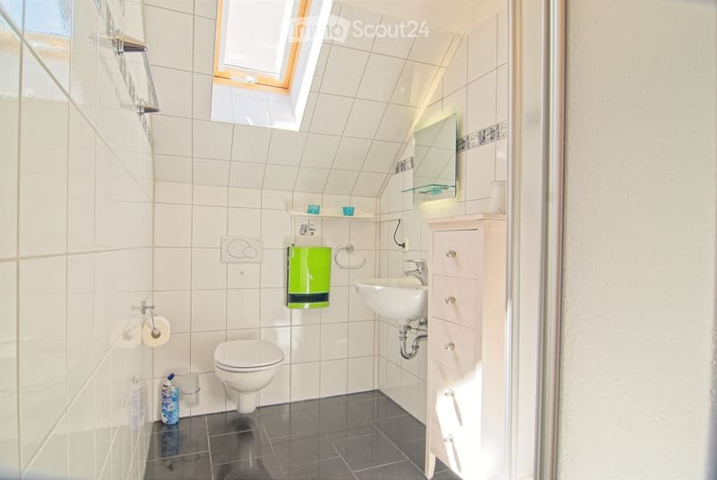 Modern bathroom with white tiles, a toilet, sink, mirror, and a small cabinet. Features a green towel, a toilet paper holder, and a small window with blinds.