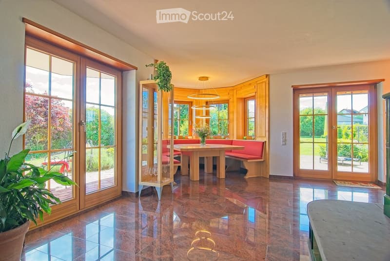 Bright, spacious room with large wooden-framed windows and doors leading to a garden. Features a central wooden table with a red bench seating, a hanging pendant light, and a potted plant near the entrance.