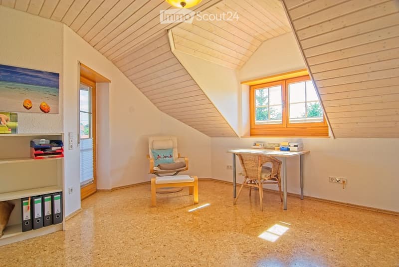 Bright, cozy attic room with wooden slanted ceilings, large windows, a comfortable chair, a small table with a wicker chair, and a shelf with books and decorative items.