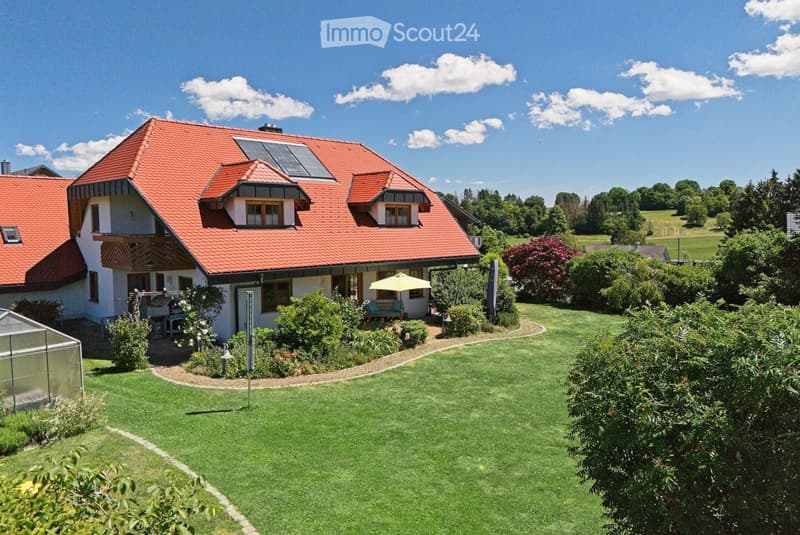Two-story house with red-tiled roof, white walls, multiple windows, and a well-maintained lawn with surrounding greenery and a patio area.