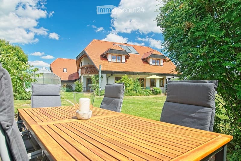 A wooden outdoor dining table with four grey chairs and a candle in the center, set in a well-maintained garden with a large house in the background.