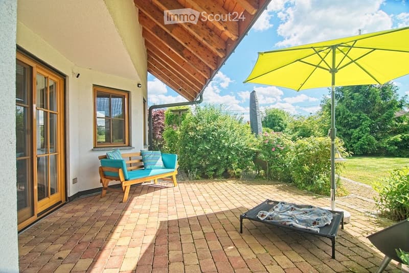 A covered patio area with a wooden bench, sun lounger, and large umbrella, surrounded by greenery and a brick-paved floor.