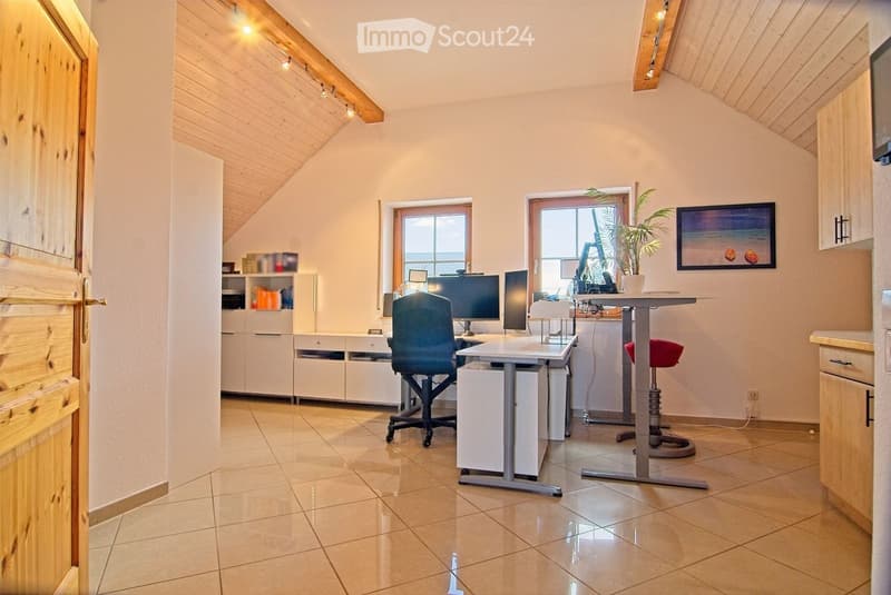 Bright, modern office space with wooden ceiling beams, tiled floor, and large windows. Features a desk with two computer screens, a comfortable chair, a small cabinet, and a potted plant on a side table.