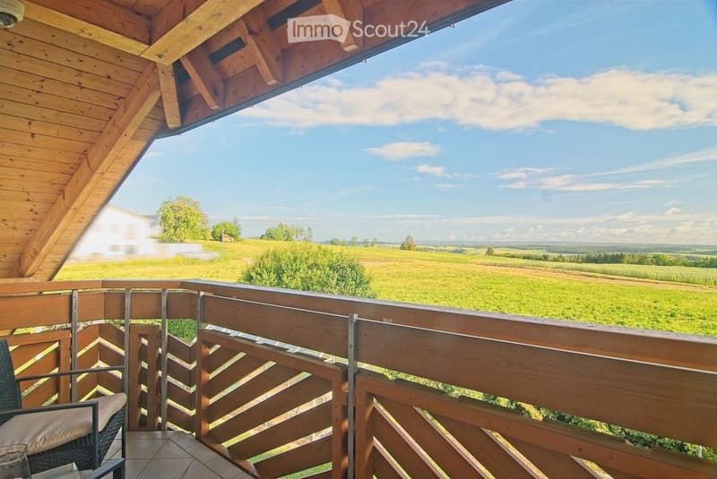 Wooden balcony with metal railing, overlooking a green field and distant landscape under a partly cloudy sky.