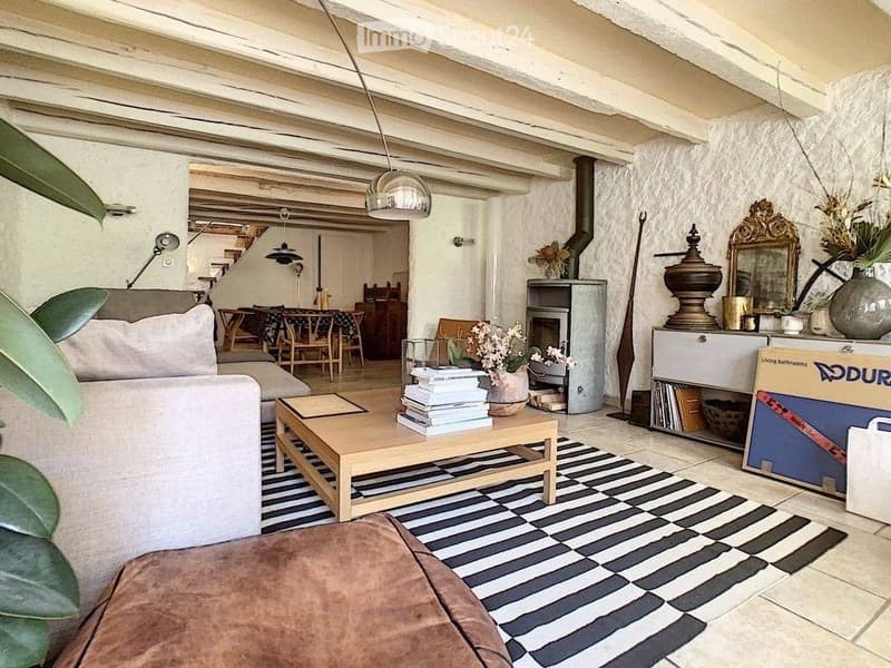 Living room with exposed wooden beams, a fireplace, a sofa, coffee table, ottoman, and dining area visible in the background.