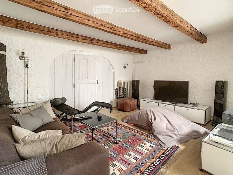 Living room with exposed wooden beams, a brown sofa with pillows, a glass coffee table, a television, a floor lamp, and a radiator.