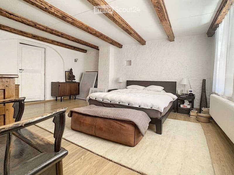 Cozy attic bedroom with wooden beams, a large bed, nightstand, chair, and a small table with a lamp.