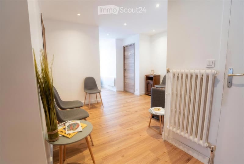 A modern, minimalist hallway with light wooden flooring, white walls, and several contemporary chairs and tables arranged neatly. A radiator is visible on the right side, and a small cabinet is placed against the wall.