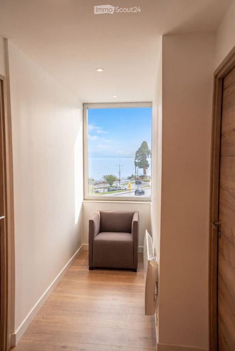 A narrow hallway with wooden flooring, a single window offering a view of the sea, and a small brown armchair placed near the window.